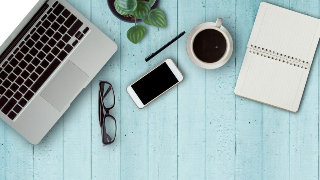 Photo of a teal desk with a laptop, phone, glasses, coffee and a plant, representing a successful mindfulness teaching career.